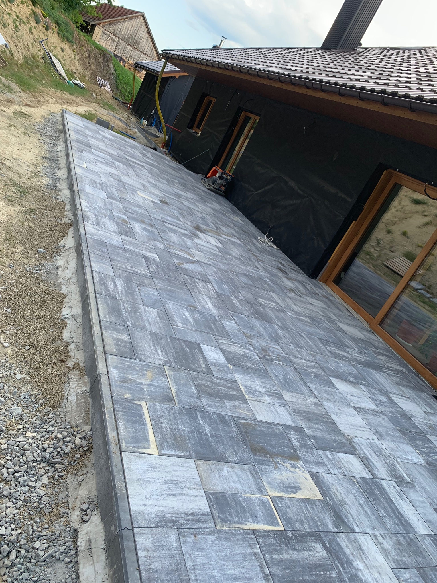 Ułożona szara kostka brukowa na posesji, widoczne obrzeża i podsypka żwirowa, w tle budynek w trakcie budowy z oknami i czarną folią elewacyjną.