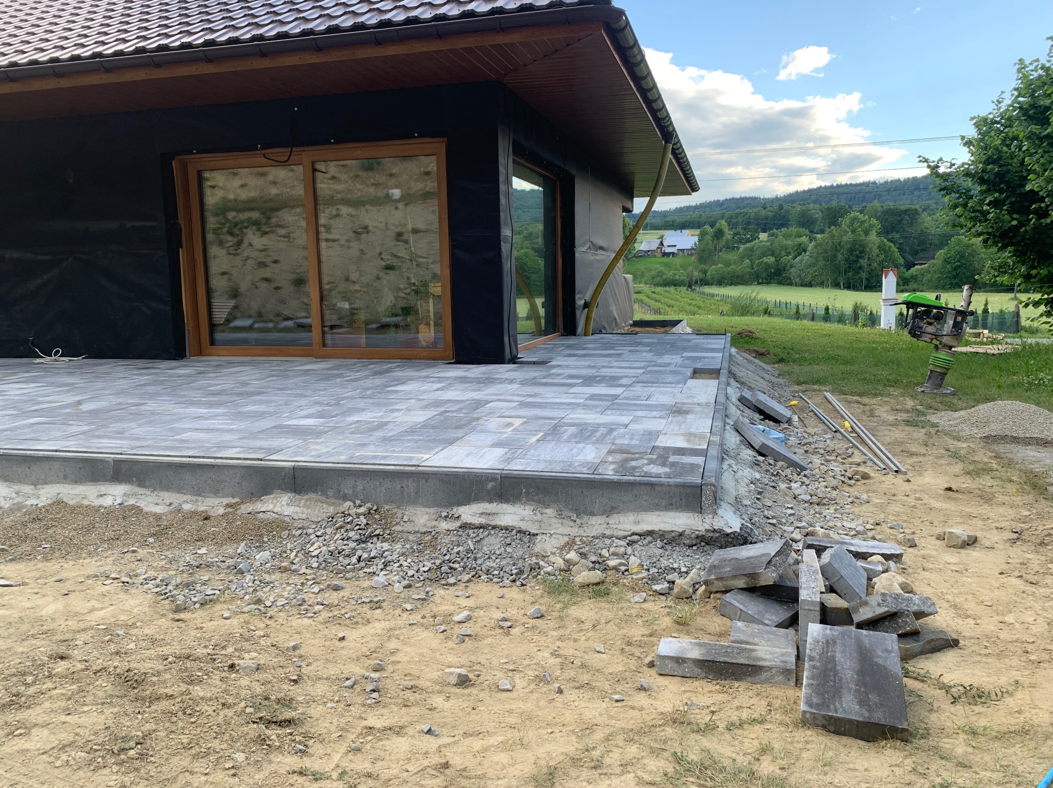 Układanie tarasu z szarej kostki brukowej przy nowo budowanym domu z widokiem na pagórkowaty krajobraz, widoczna ubijarka w tle.