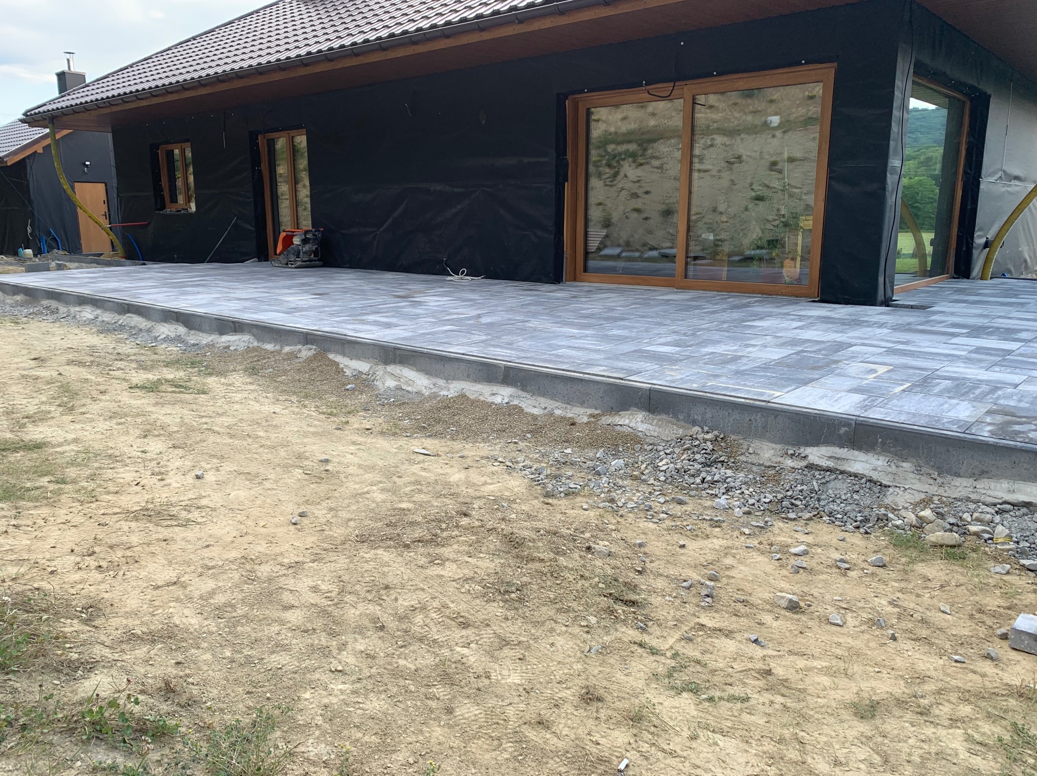 Nowoczesny taras z szarych płyt kamiennych w trakcie budowy, widoczny zagęszczarka wibracyjna, dom w stanie surowym zamkniętym w tle.