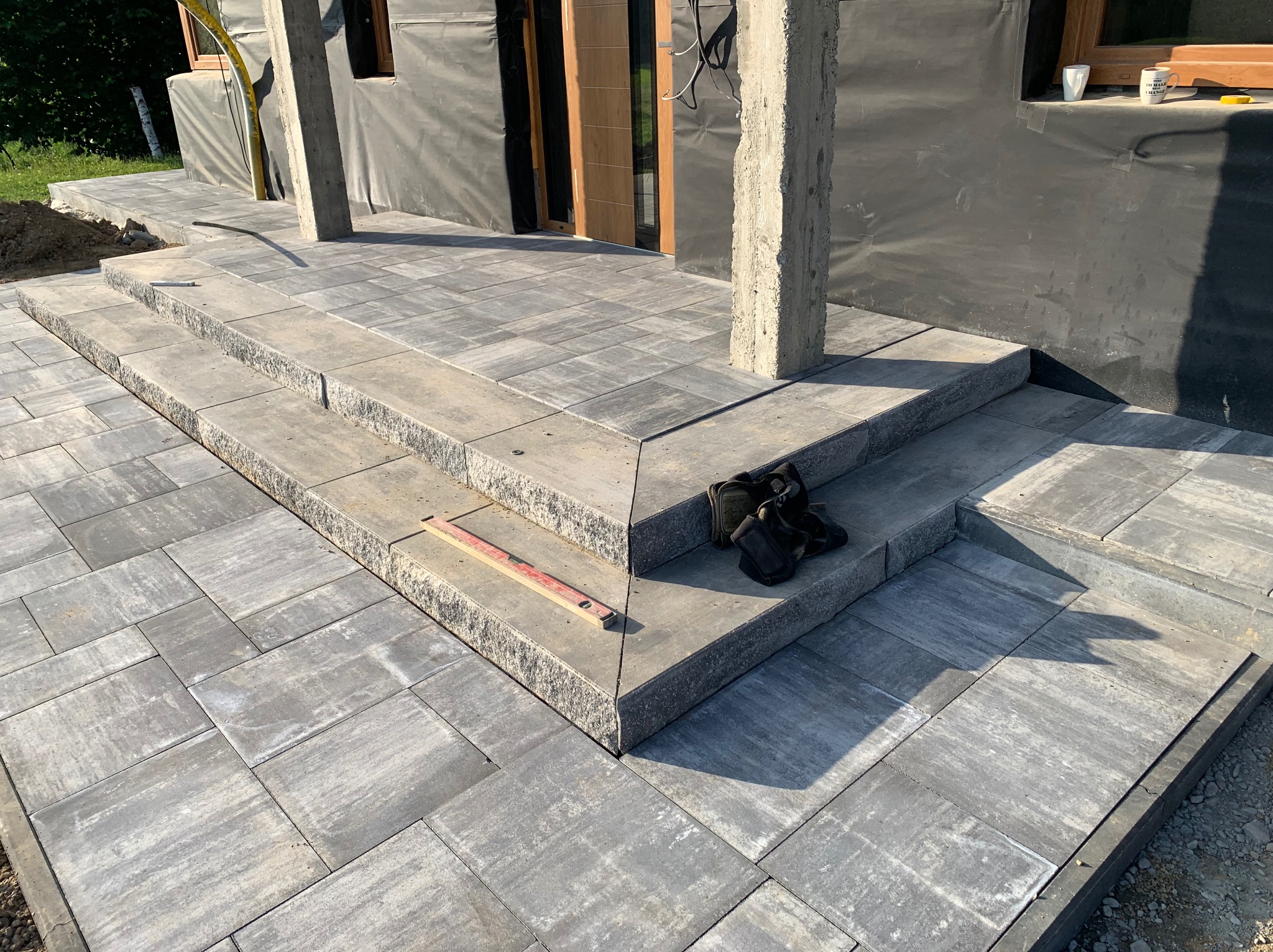 Schody wejściowe z szarych płyt kamiennych, częściowo wykończone, z poziomnicą i rękawicami roboczymi na jednym ze stopni, widoczne elementy elewacji budynku w trakcie prac.