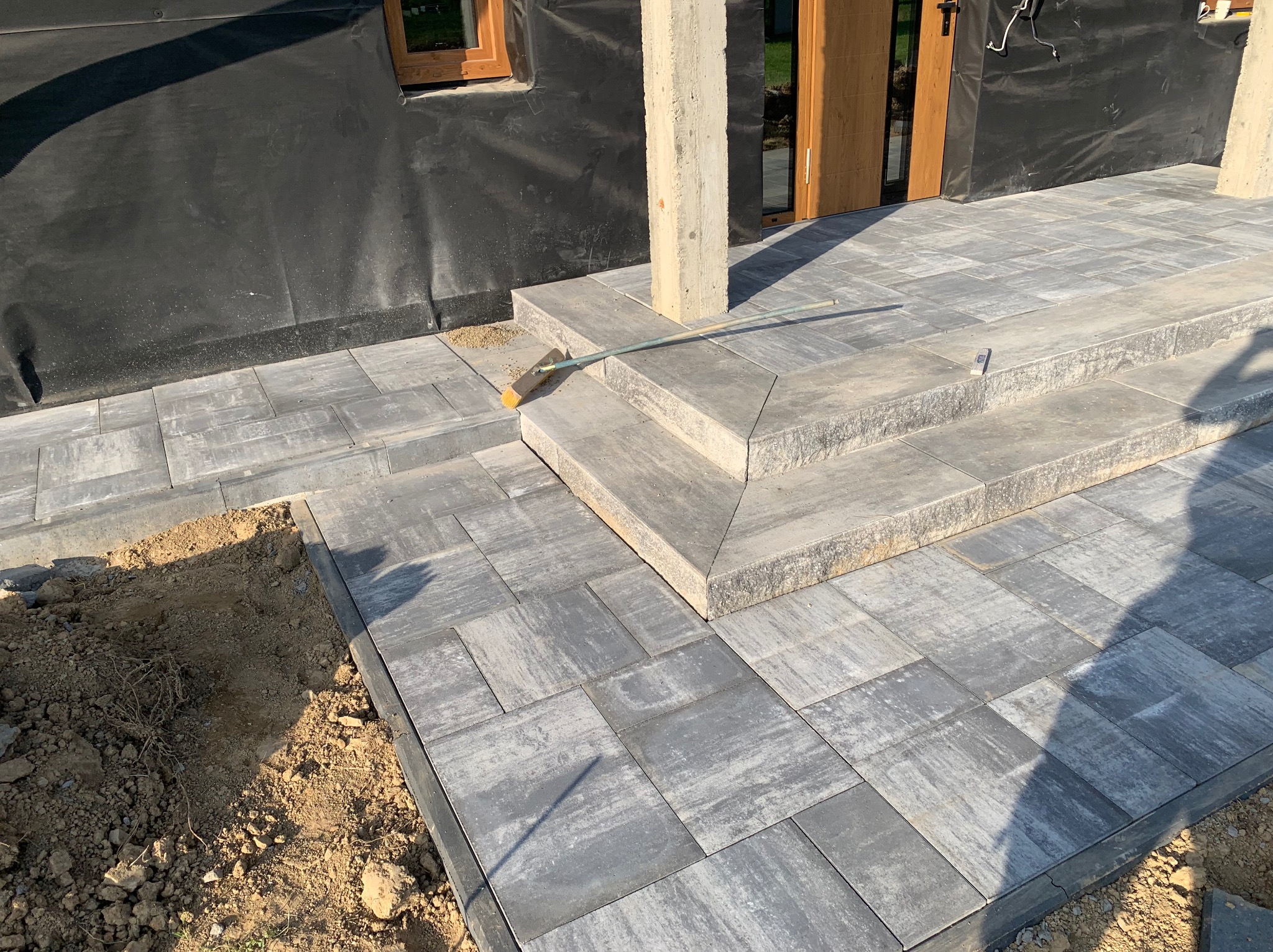 Nowo ułożona, szara kostka brukowa tworząca taras i schody wejściowe do domu w trakcie wykańczania, widoczny betonowy filar i czarna folia na ścianie budynku, z boku odsłonięta ziemia, na schodach...