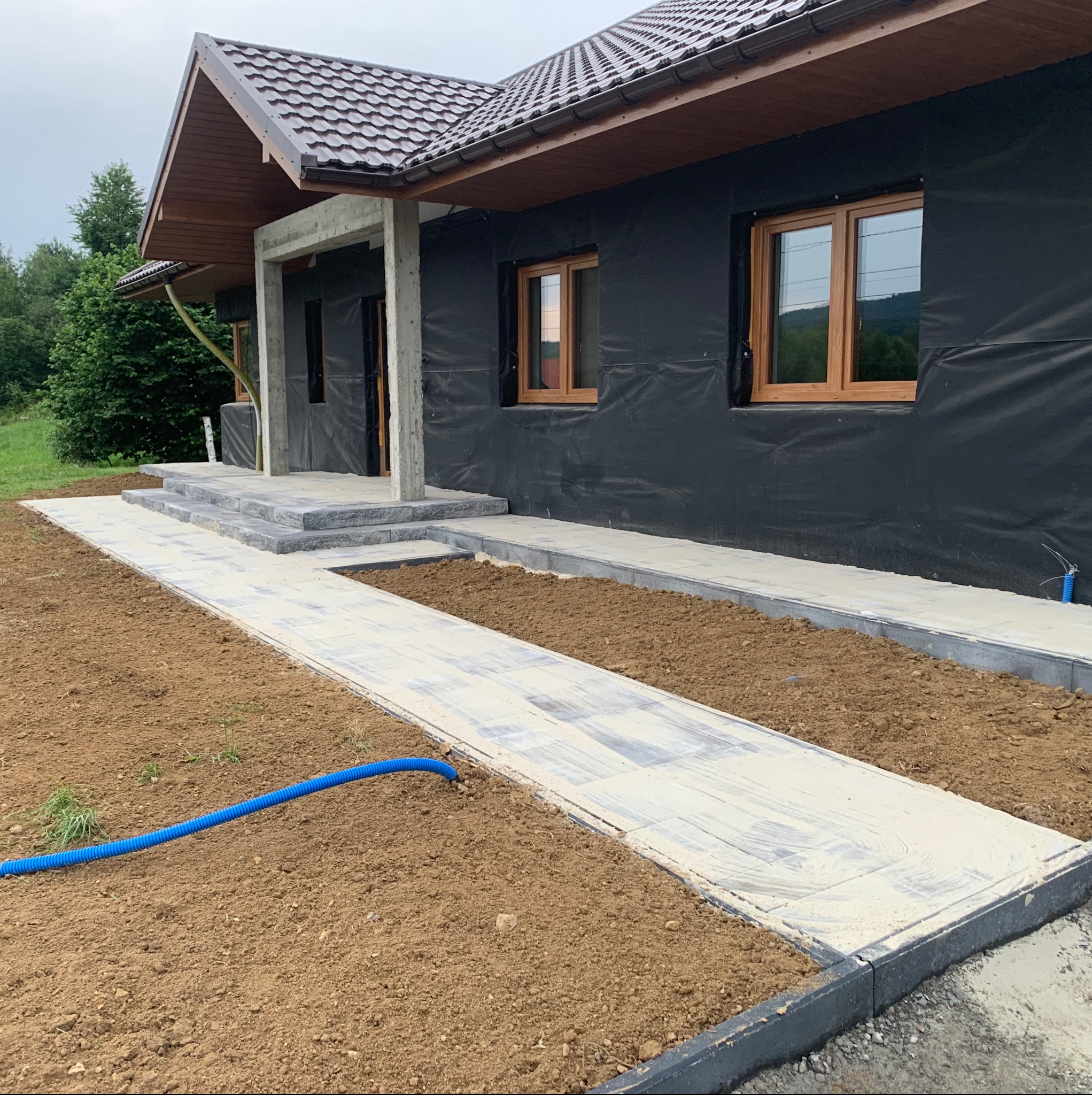 Nowo ułożony, jasnoszary chodnik z kostki brukowej prowadzący do domu w trakcie budowy, widoczny niebieski karbowany przewód na przygotowanym podłożu.