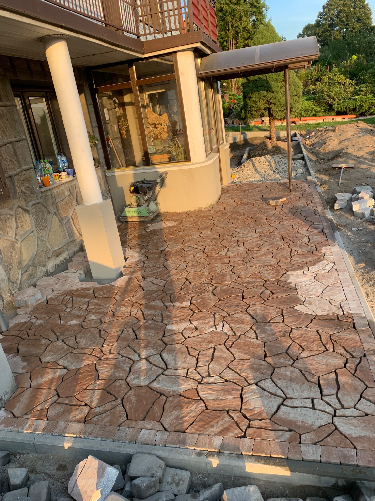 Częściowo ułożony chodnik z nieregularnych, brązowo-beżowych kamieni, obramowany szarym obrzeżem, widoczny w trakcie prac wykończeniowych przy domu z kamienną elewacją i kolumną.