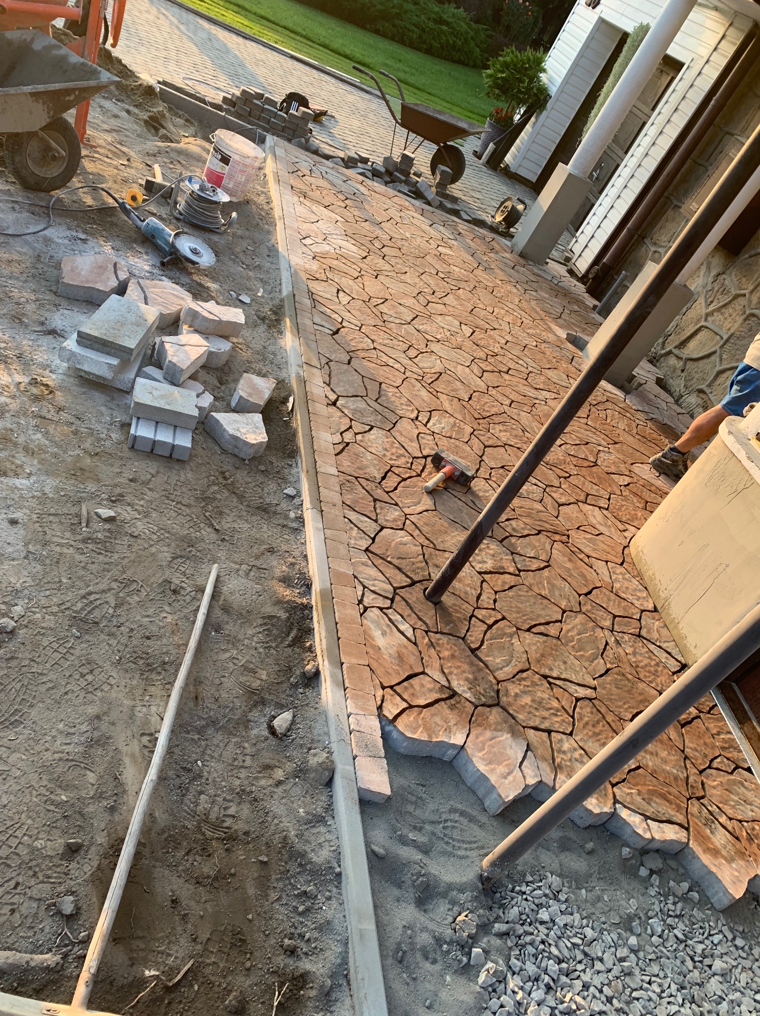 Układanie nieregularnej kostki brukowej w odcieniach brązu i beżu na podjeździe, widoczne narzędzia pracy, wózek, stos kostki, obrzeża i częściowo ułożona nawierzchnia.