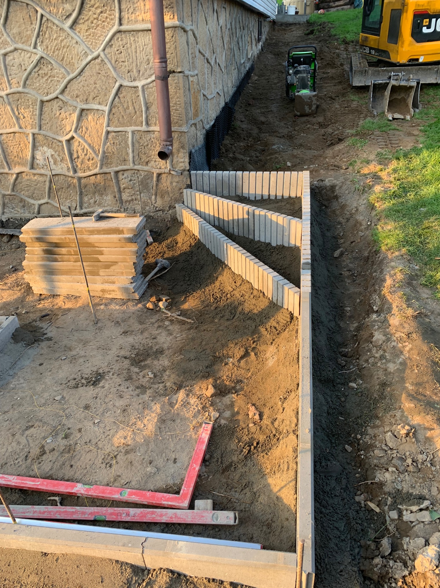 Budowa schodów terenowych z betonowych elementów, widoczne narzędzia pomiarowe, wykop ziemny i zagęszczarka w tle.