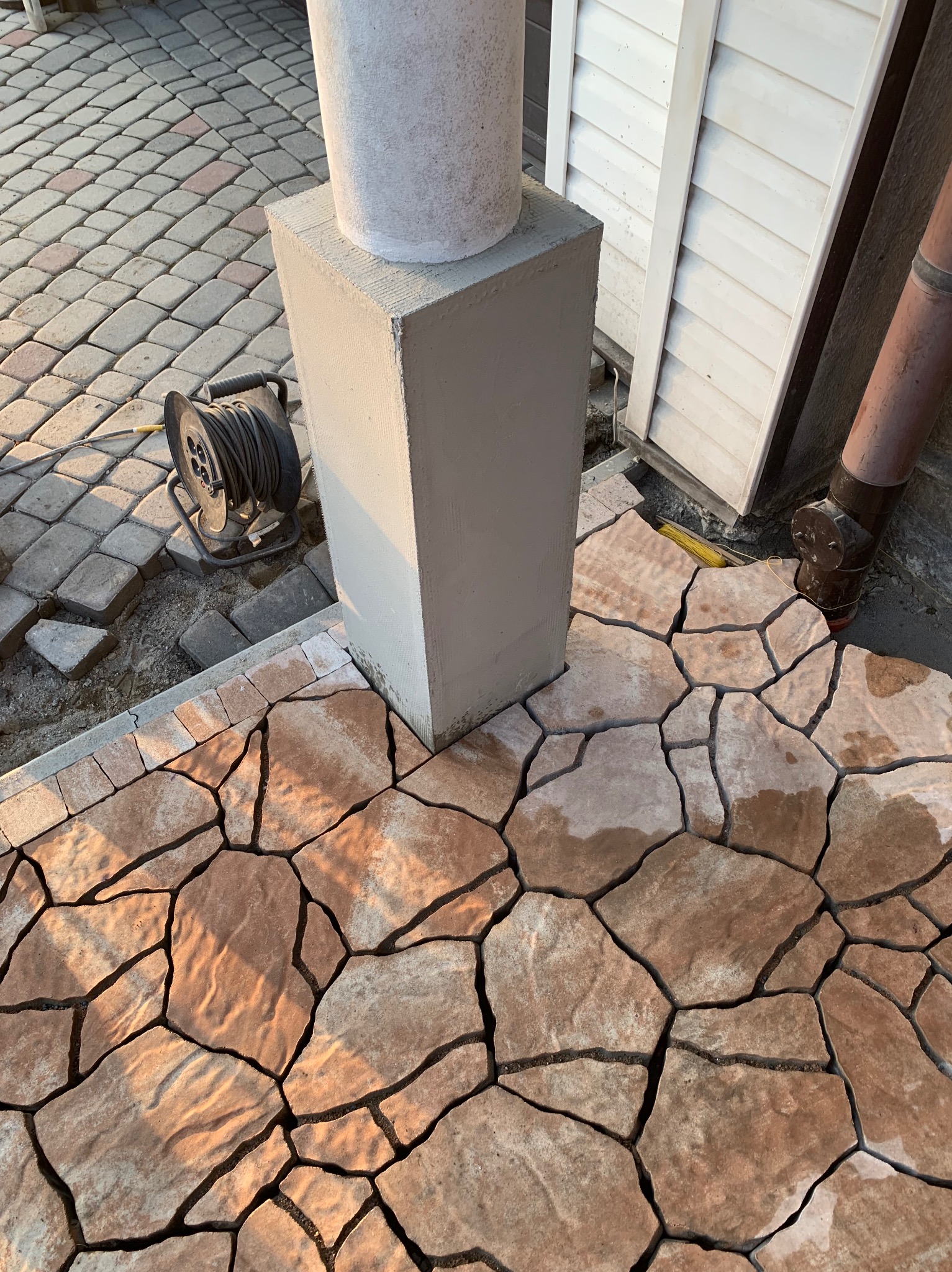 Fragment układanej nawierzchni z nieregularnych płyt kamiennych w odcieniach brązu, sąsiadujących z kostką brukową i betonowym słupkiem, widoczna zwinięta przedłużka elektryczna.