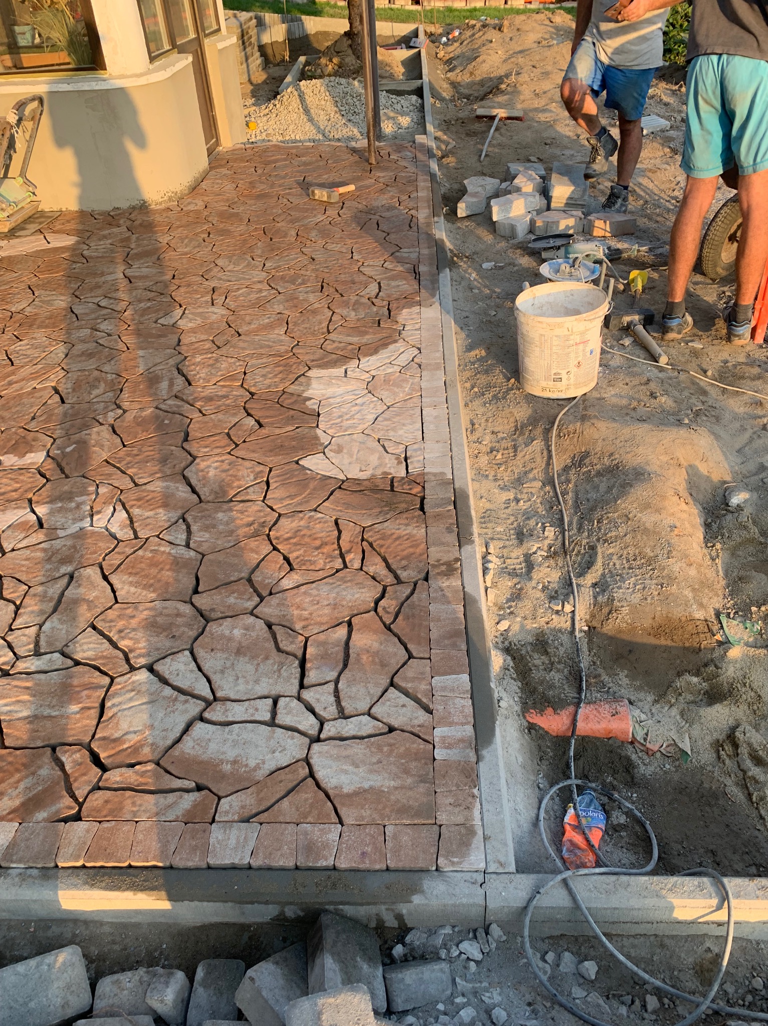 Nieregularny wzór z brązowych kamieni tworzy ścieżkę, obrzeże z małych kostek, widoczne narzędzia i materiały budowlane, dwóch pracowników w tle.