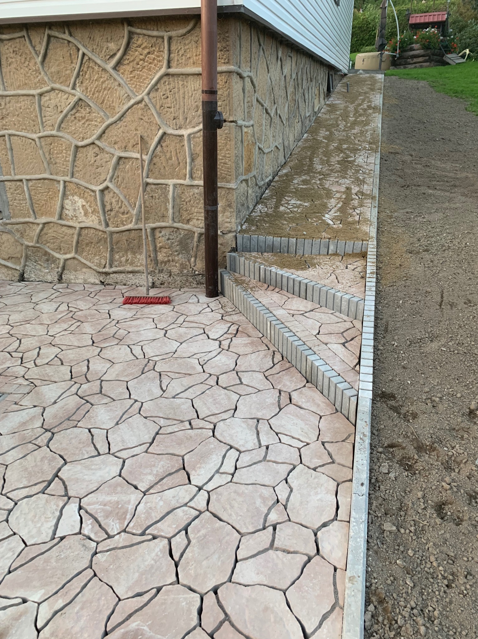 Jasna, nieregularna kostka na posesji z kamienną ścianą budynku w tle, widoczna rynna i szczotka do zamiatania.