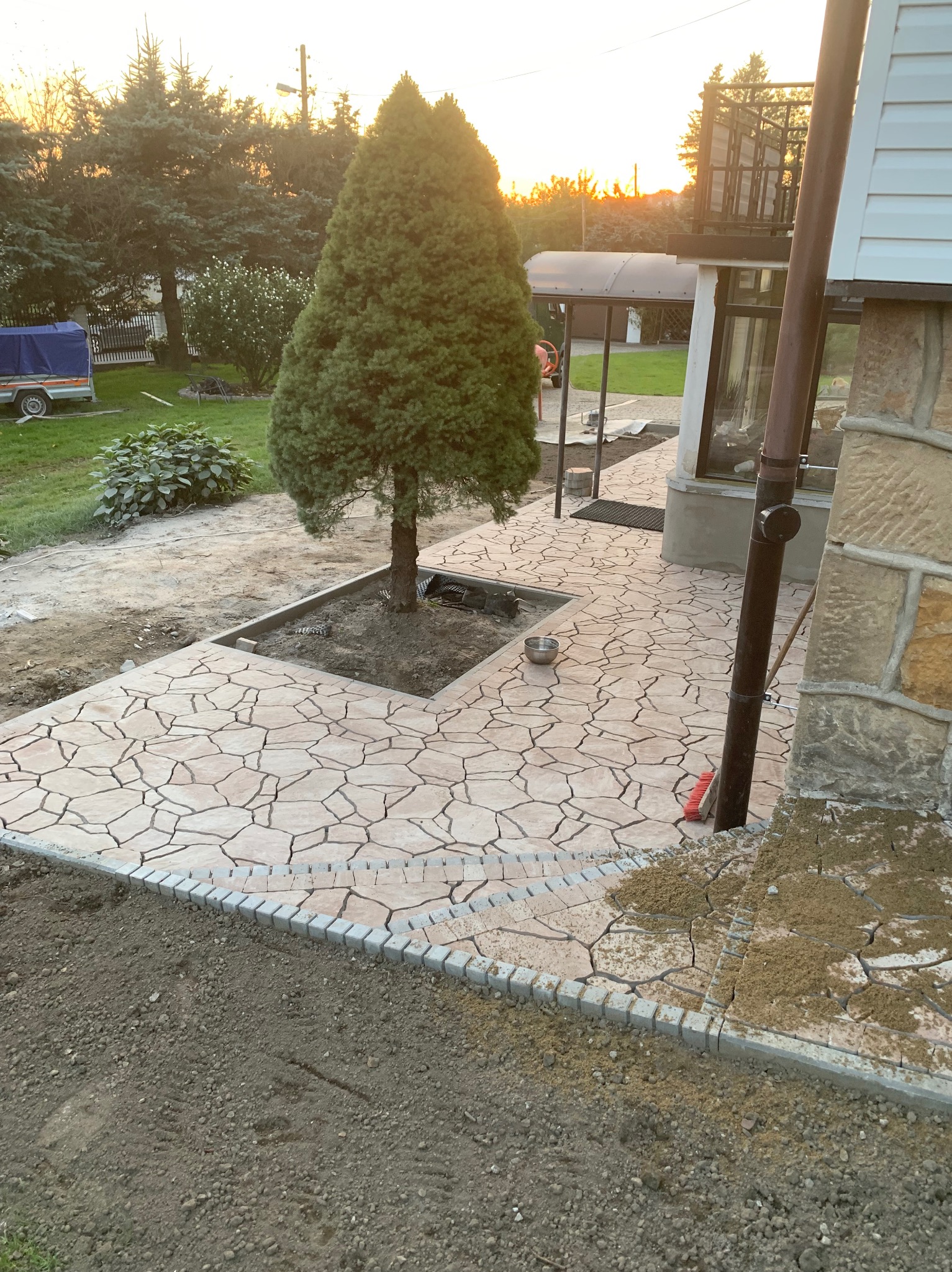 Kremowa, nieregularna nawierzchnia z kamienia ułożona wokół drzewa iglastego, obrzeża z szarej kostki, widoczny fragment elewacji domu z kamienną okładziną, w tle zabudowa i zachodzące słońce.