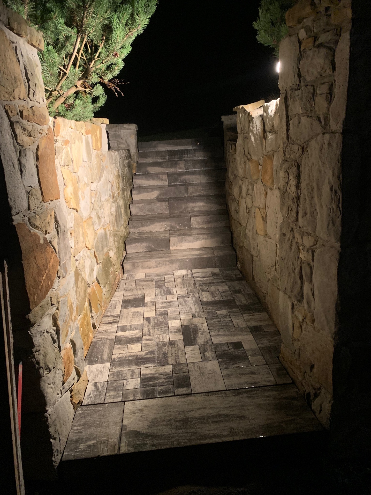 Kamienne schody prowadzące w górę, oświetlone punktowym światłem, otoczone murem z nieregularnych kamieni, z fragmentem iglastego drzewa w rogu.