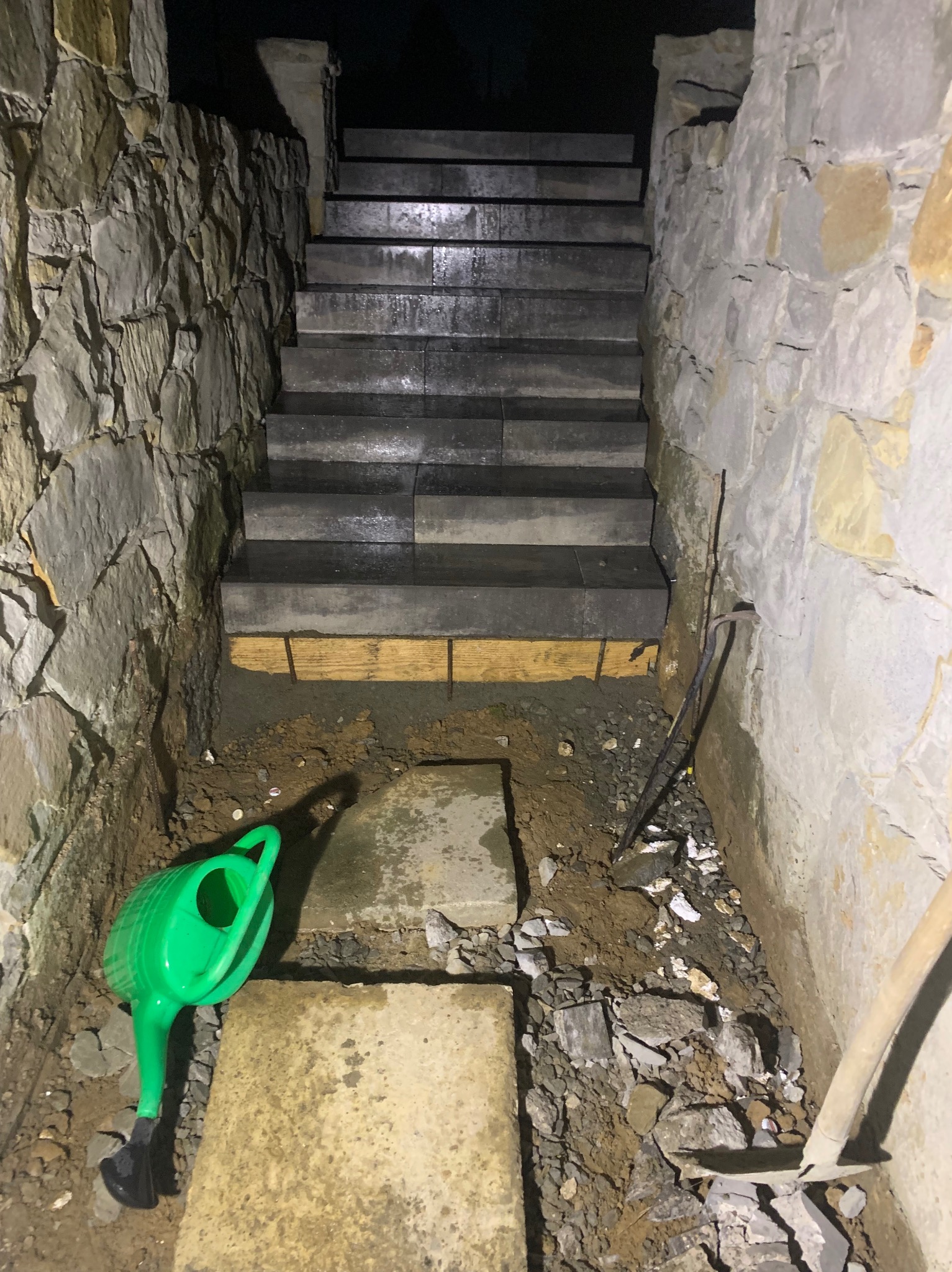 Budowa kamiennych schodów zewnętrznych w trakcie realizacji, widoczne narzędzia, zielona konewka i fragment ściany z kamienia łupanego.