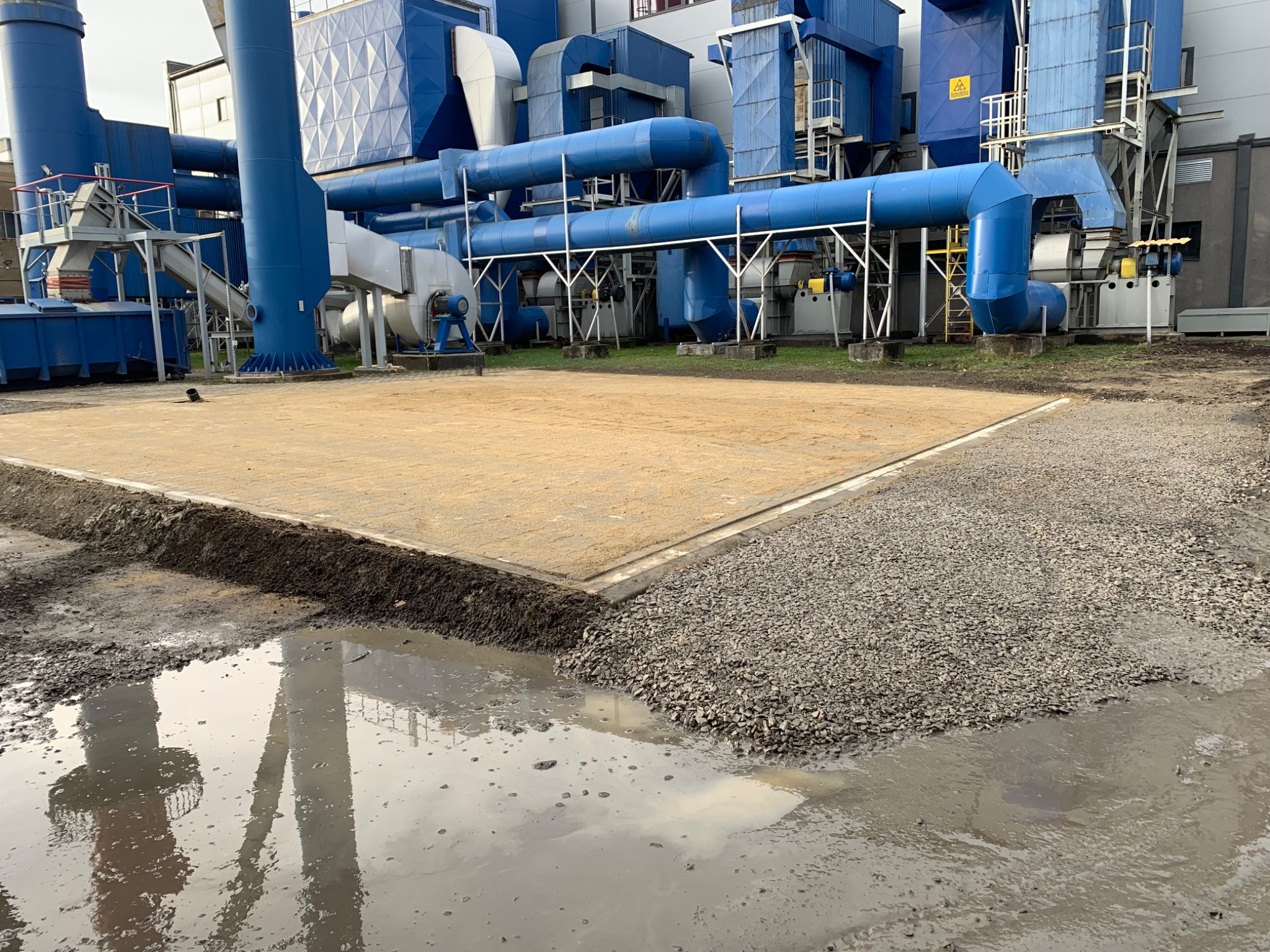 Przygotowanie terenu pod budowę: wyrównany piasek, obok warstwa gruzu, kałuża z odbiciem przemysłowej instalacji w tle.