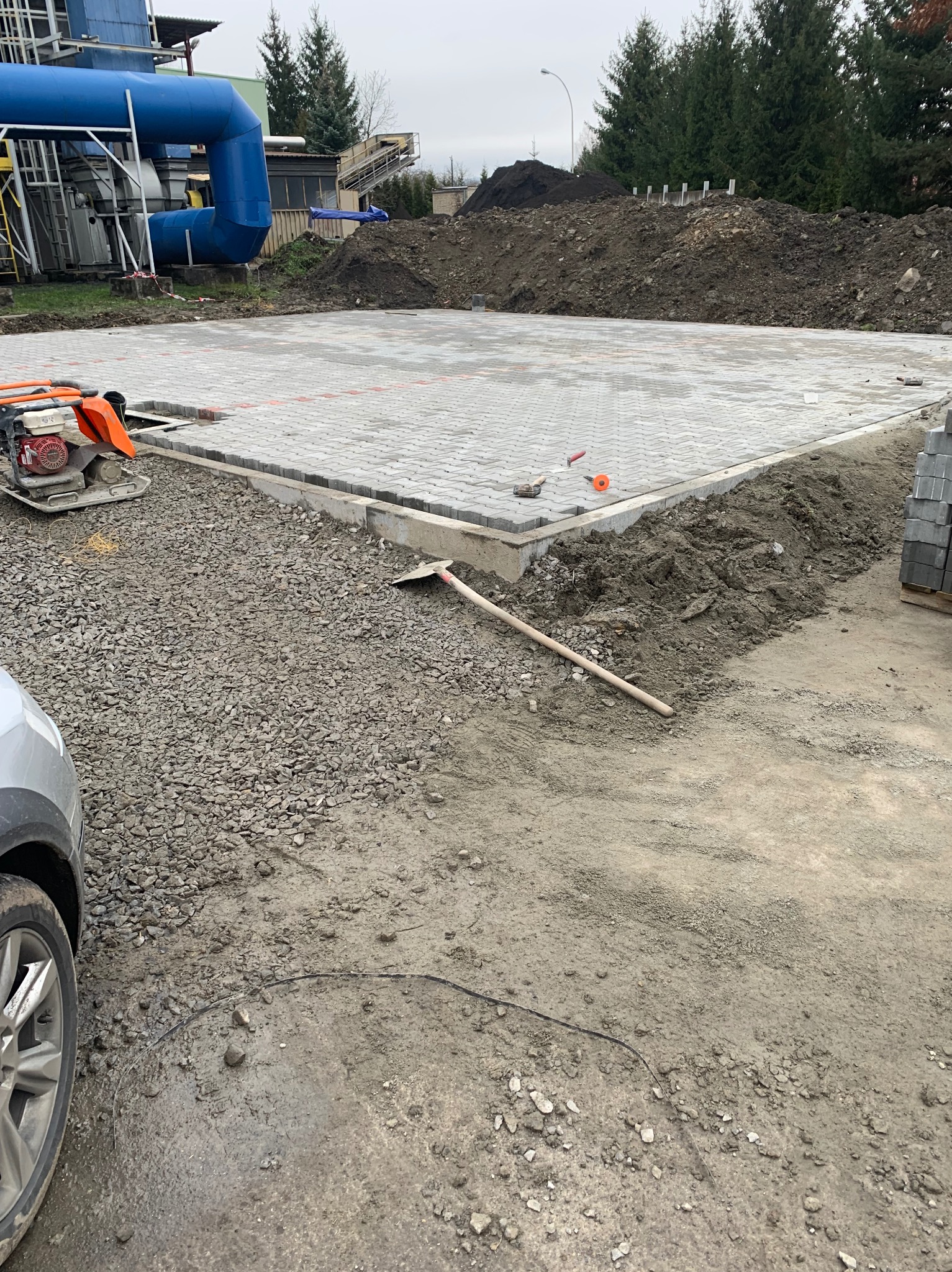 Przygotowany teren pod plac z częściowo ułożoną szarą kostką, widoczna ubijarka, szpadel, hałda ziemi w tle oraz fragment samochodu.