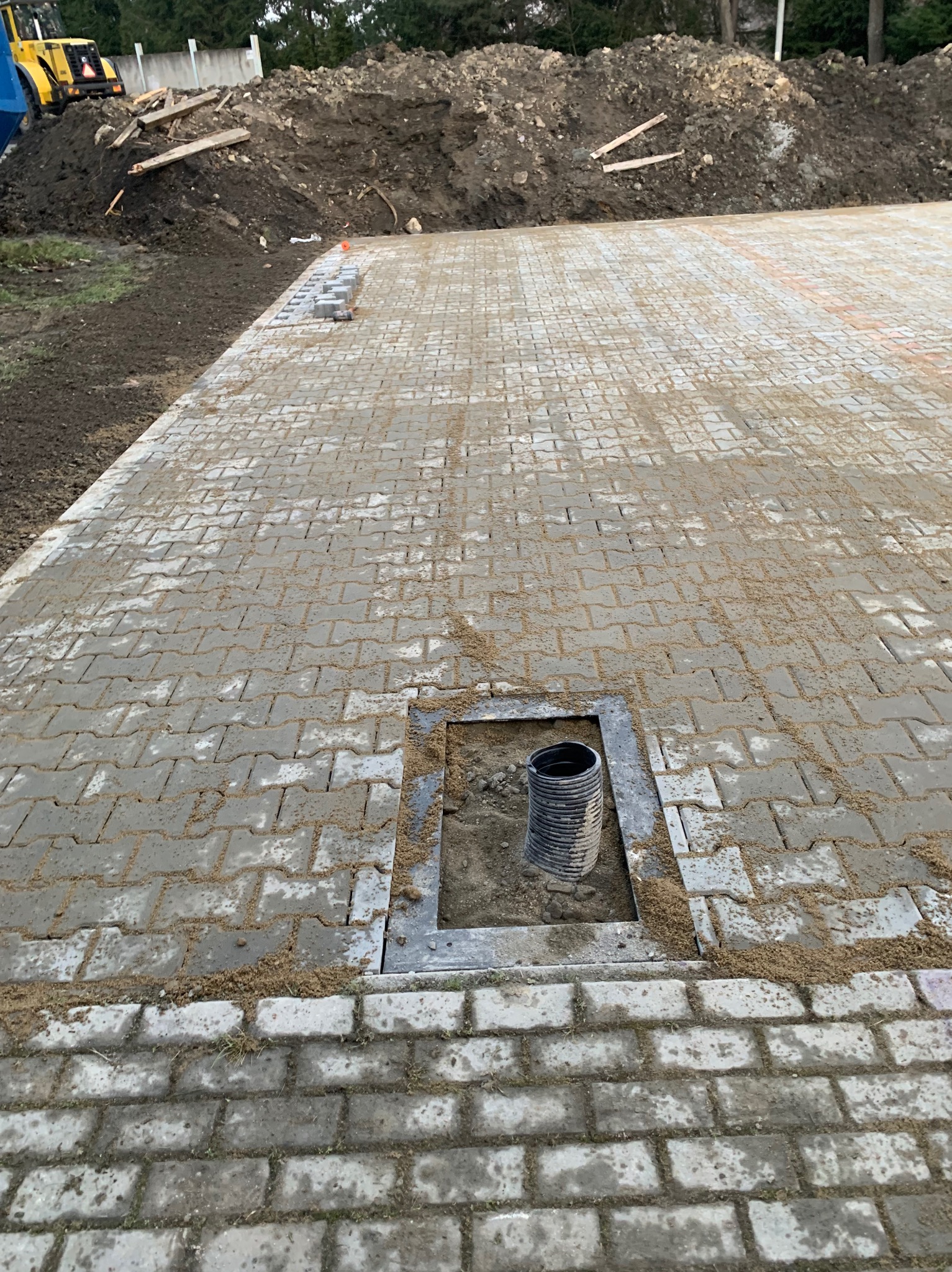 Częściowo ułożona szara kostka brukowa w Żyznowie, z widocznym otworem i rurą kanalizacyjną. W tle hałda ziemi i sprzęt budowlany.