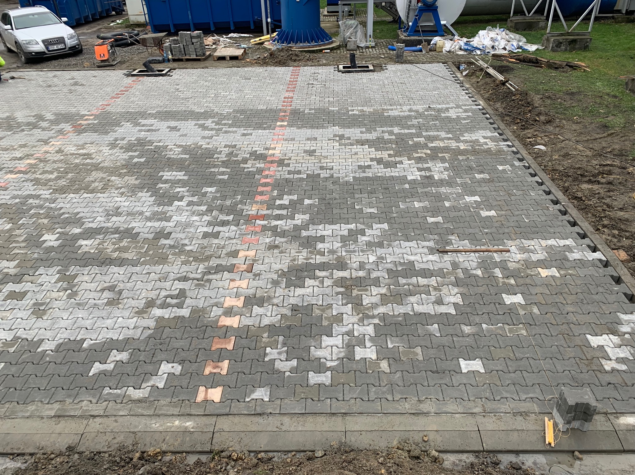 Ułożona szara kostka brukowa typu 'psia kość' z pomarańczowymi akcentami, widoczne studzienki kanalizacyjne i zaparkowany samochód w tle, teren budowy.
