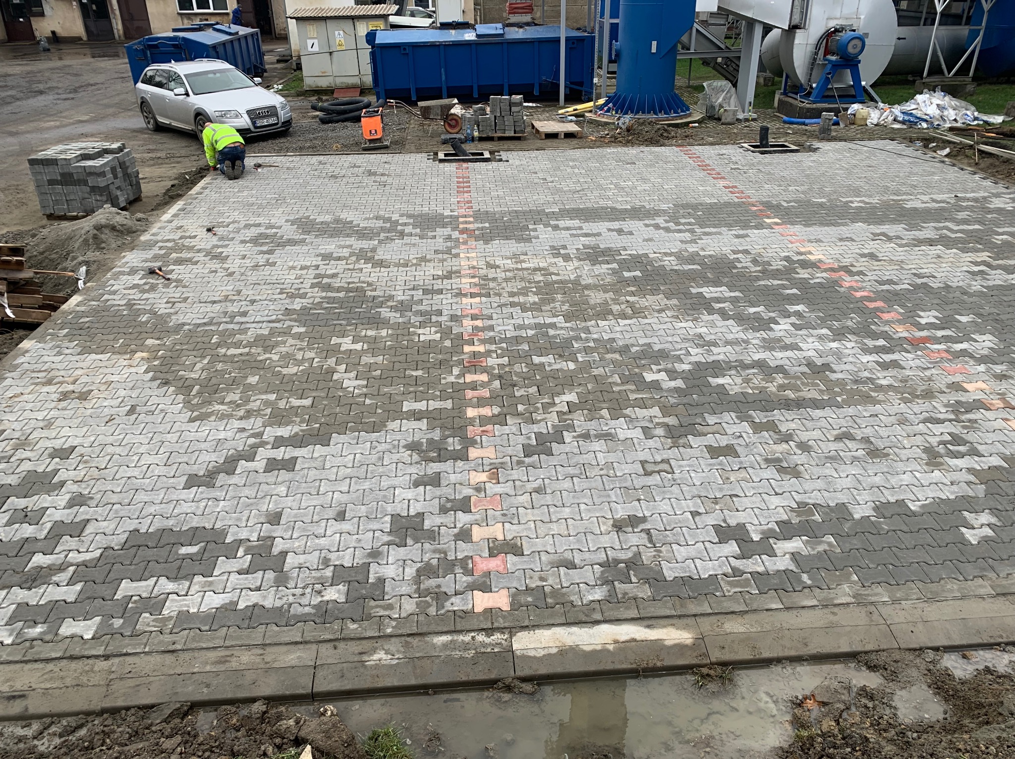 Prace brukarskie w toku: układanie szarej kostki betonowej w jodełkę z rzędem ceglastych akcentów, widoczny pracownik w odblaskowej kamizelce, stos kostki, narzędzia i maszyny budowlane w tle.