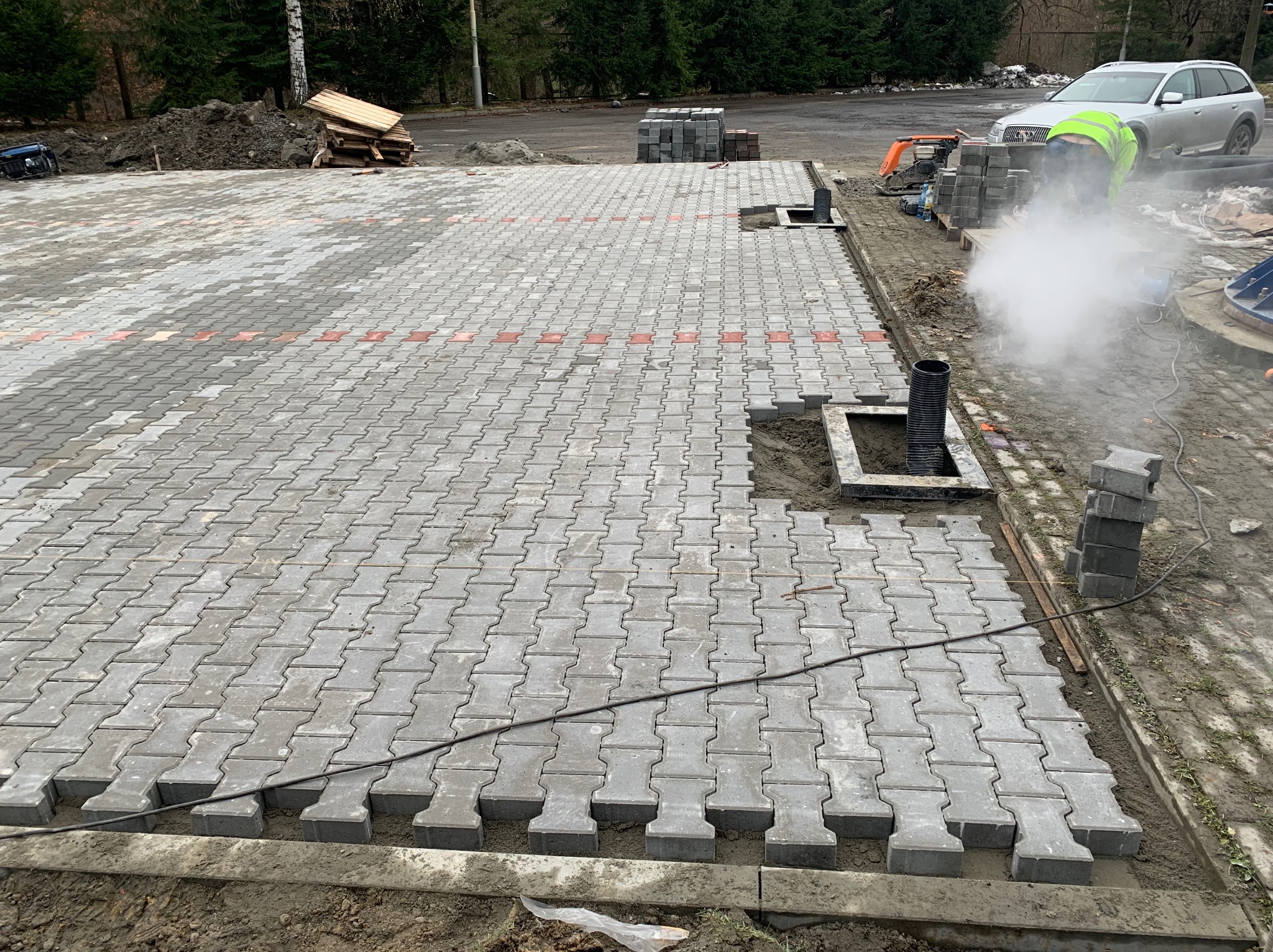 Fragment placu z ułożoną szarą kostką brukową typu I, widoczny pracownik w odblaskowej kamizelce, palety z kostką w tle, oraz rury kanalizacyjne w przygotowanych otworach.