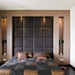 Sypialnia w odcieniach brązu i czerni z tapicerowanym wezgłowiem łóżka w formie kwadratów, oświetlonymi wnękami po bokach i obrazem na ścianie.