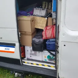 Otwarty tył białego vana załadowanego kartonami, torbami i paczkami, w tym pudełka z odkurzaczem Sencor i maszyną do szycia Singer, widoczne trawa i hak holowniczy.