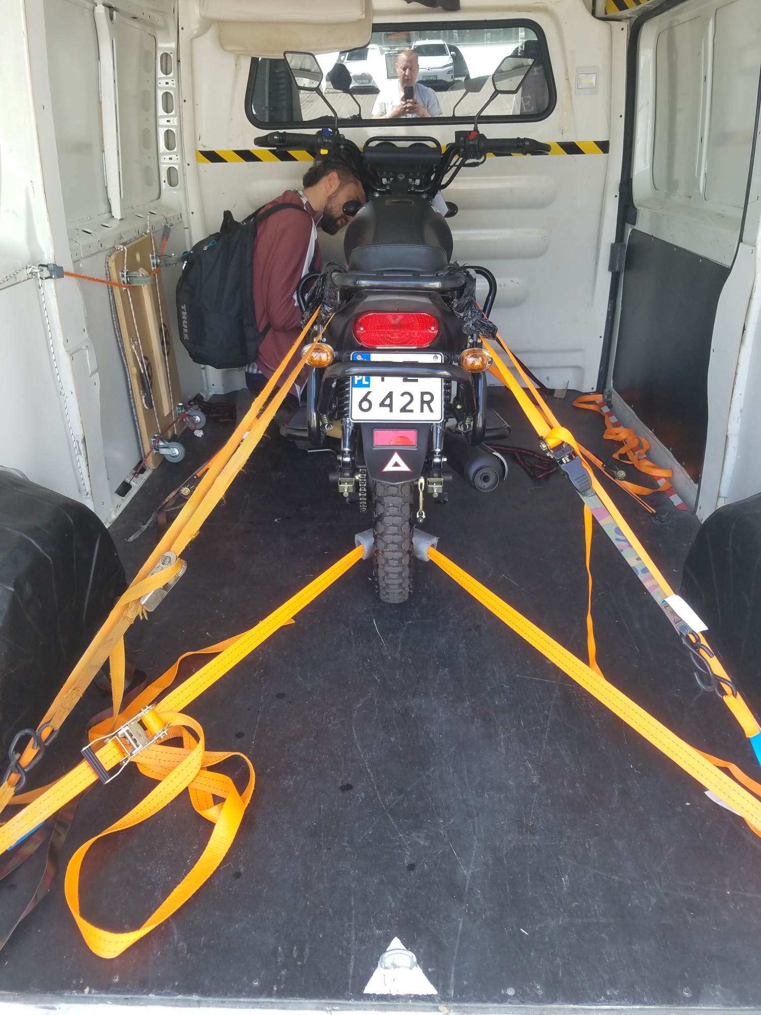 Motocykl zabezpieczony pomarańczowymi pasami transportowymi wewnątrz białej furgonetki, widoczny mężczyzna z plecakiem sprawdzający mocowanie, w tle odbicie drugiego mężczyzny robiącego zdjęcie...