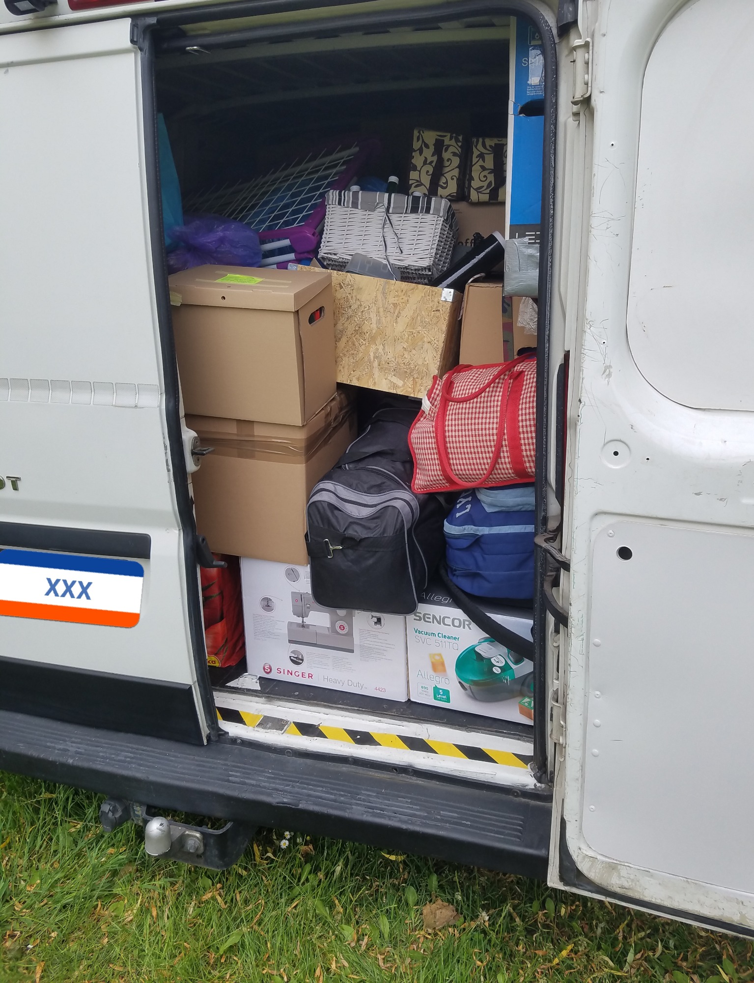 Otwarty tył białego vana załadowanego kartonami, torbami i paczkami, w tym pudełka z odkurzaczem Sencor i maszyną do szycia Singer, widoczne trawa i hak holowniczy.