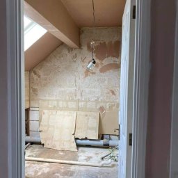 racsobuild - Remontowane poddasze z oknem dachowym: ściany po skrobaniu, przygotowane do szpachlowania, widoczne rury i prowizoryczne oświetlenie na kablu, widok zza otwartych drzwi.