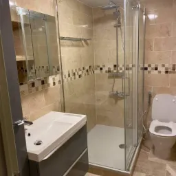 Wykończona łazienka z beżowymi płytkami, kabiną prysznicową ze szklanymi drzwiami, umywalką z szafką w kolorze szarym oraz toaletą z bidetką.