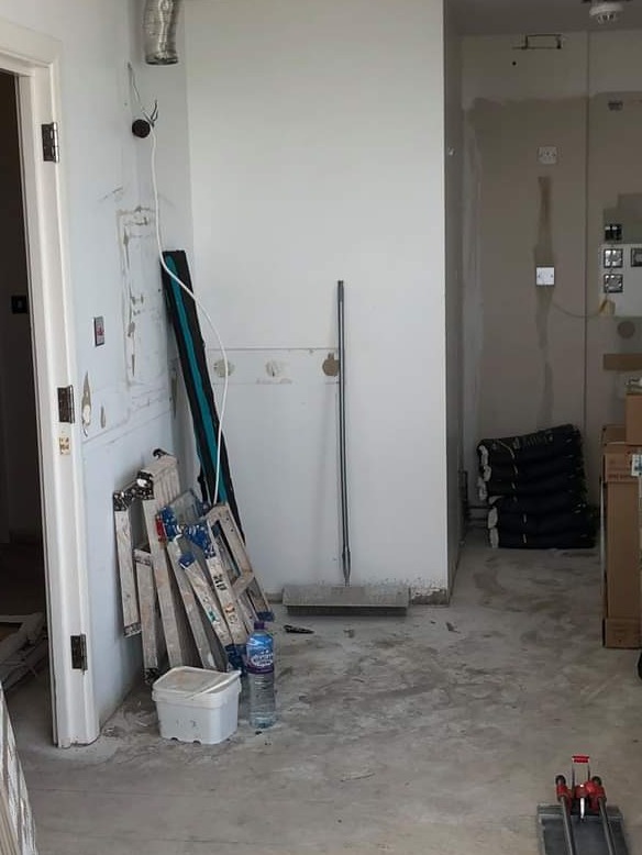 Wnętrze w trakcie prac przygotowawczych: surowe ściany, goła podłoga, drabiny oparta o ścianę, wiadro z pokrywką, butelka wody i narzędzia do cięcia płytek na podłodze.