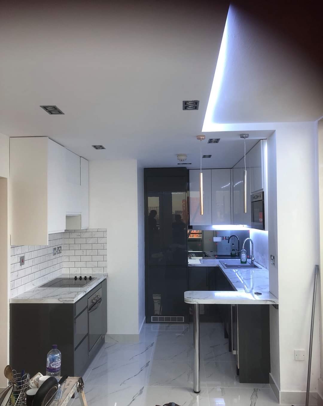 Nowoczesna kuchnia z białymi i ciemnoszarymi szafkami, marmurowym blatem i podłogą, oświetleniem LED podszafkowym i wiszącymi lampami, widok z poziomu podłogi z elementami drabiny malarskiej.