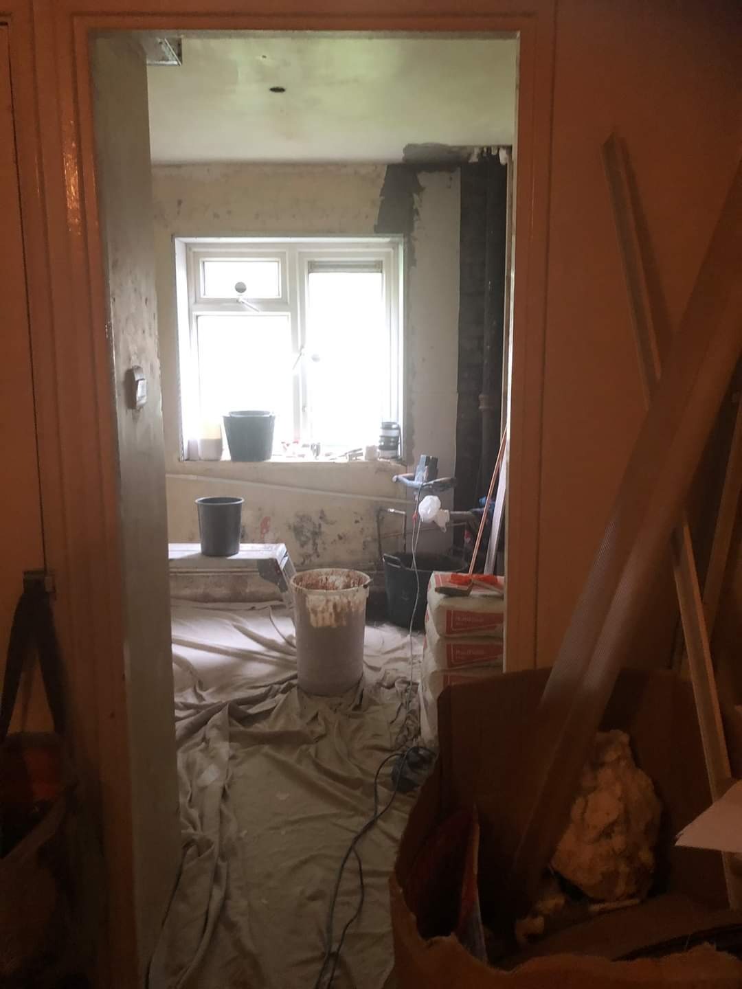 Wnętrze pokoju w trakcie prac przygotowawczych do wykończenia, widok z sąsiedniego pomieszczenia, podłoga zabezpieczona folią malarską, widoczne wiadra, szpachla i materiały budowlane.