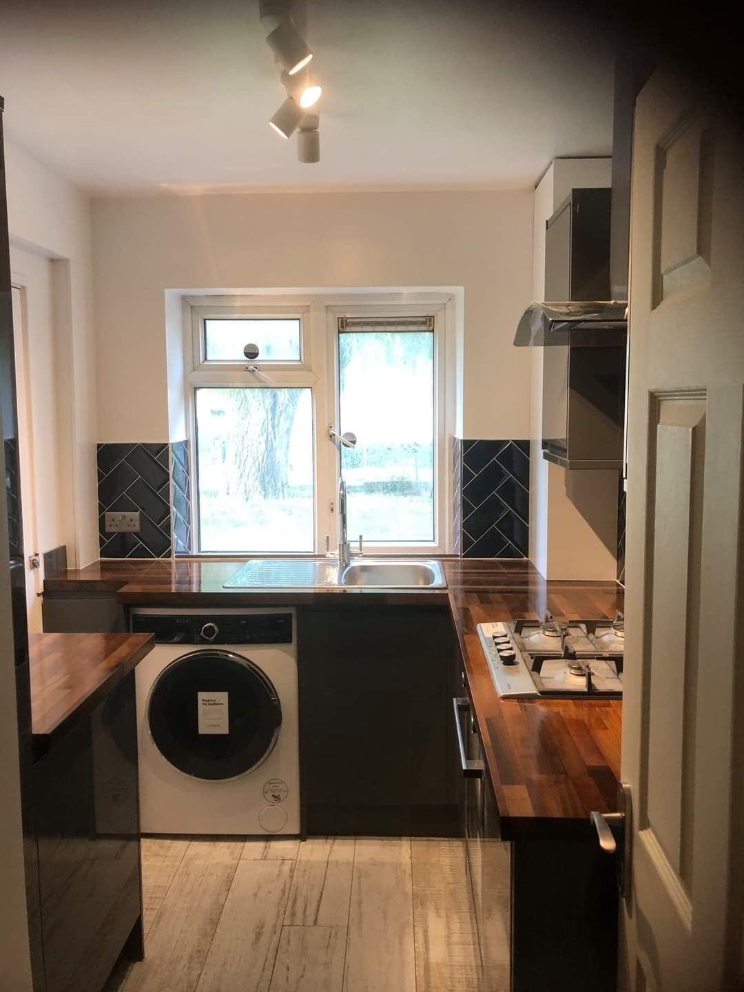 Wykończona kuchnia z ciemnymi szafkami, drewnianym blatem, czarnymi płytkami w jodełkę nad blatem i pralką w zabudowie pod oknem.