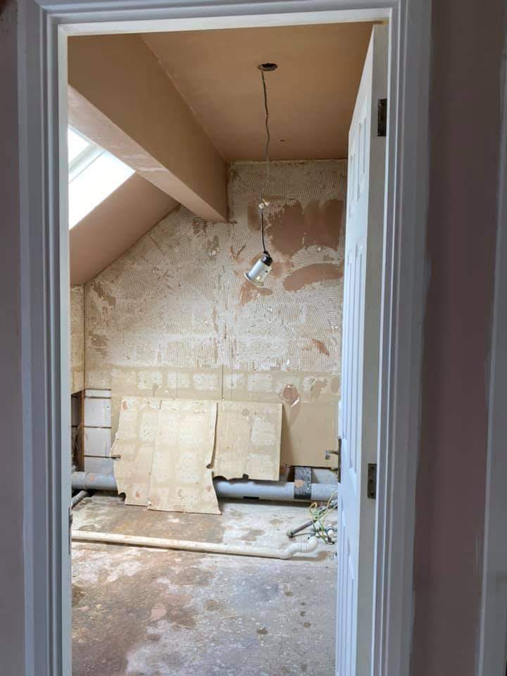 Remontowane poddasze z oknem dachowym: ściany po skrobaniu, przygotowane do szpachlowania, widoczne rury i prowizoryczne oświetlenie na kablu, widok zza otwartych drzwi.