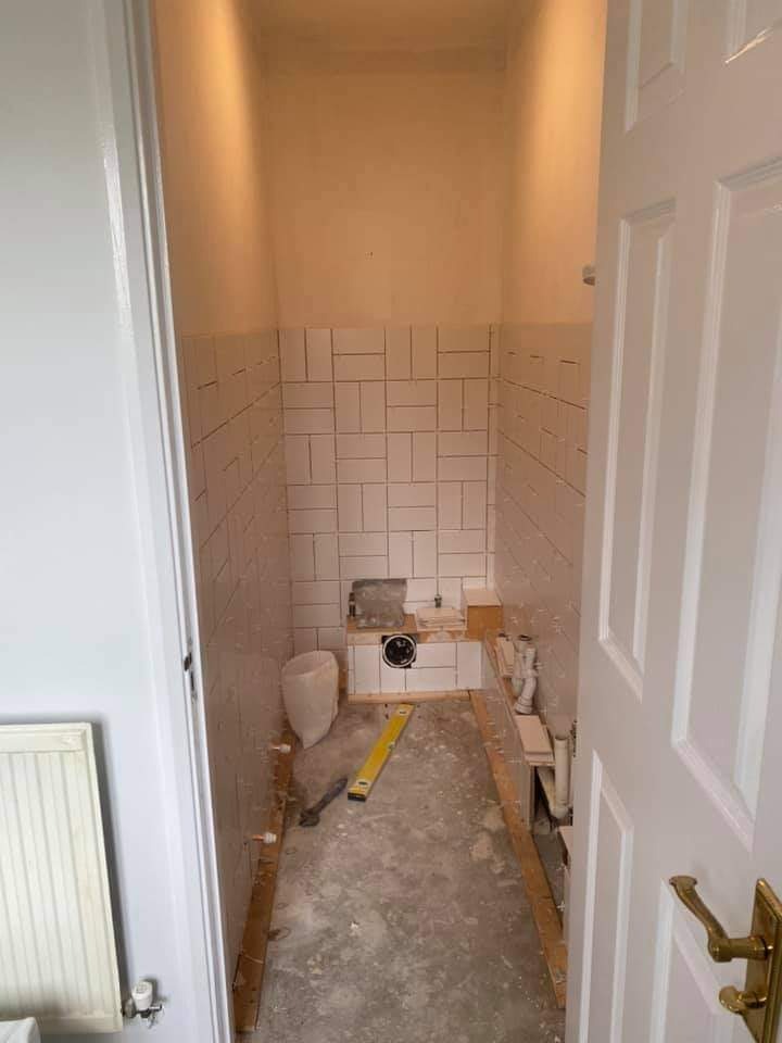 Mała łazienka w trakcie remontu: białe, prostokątne płytki ułożone w cegiełkę na ścianach do połowy wysokości, surowy beton na podłodze, poziomica, biała umywalka z odpływem, białe drzwi z mosiężną...