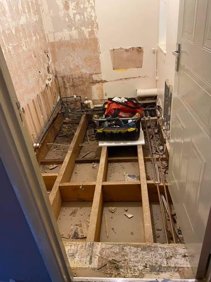 Łazienka w trakcie demontażu, widoczne odsłonięte rury, drewniana konstrukcja pod podłogą, ściany częściowo obdarte z płytek i resztek zaprawy, skrzynka z narzędziami na podwyższeniu.