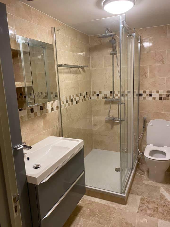 Wykończona łazienka z beżowymi płytkami, kabiną prysznicową ze szklanymi drzwiami, umywalką z szafką w kolorze szarym oraz toaletą z bidetką.