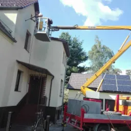 Pracownik na podnośniku koszowym czyści elewację domu jednorodzinnego za pomocą myjki ciśnieniowej; widoczny fragment dachu z dachówki ceramicznej i instalacją fotowoltaiczną na sąsiednim budynku.