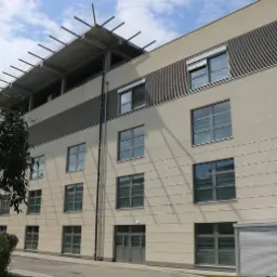 Zewnętrzny widok nowo wybudowanego, trzypiętrowego budynku szpitala z płaskim dachem i szarą stolarką okienną, częściowo przysłoniętego zielenią drzew.