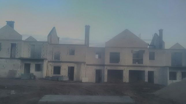 Surowy stan budynku w Dusznikach, widoczne ściany z pustaków, otwory okienne i kominy w trakcie budowy, teren wokół przygotowany pod dalsze prace.