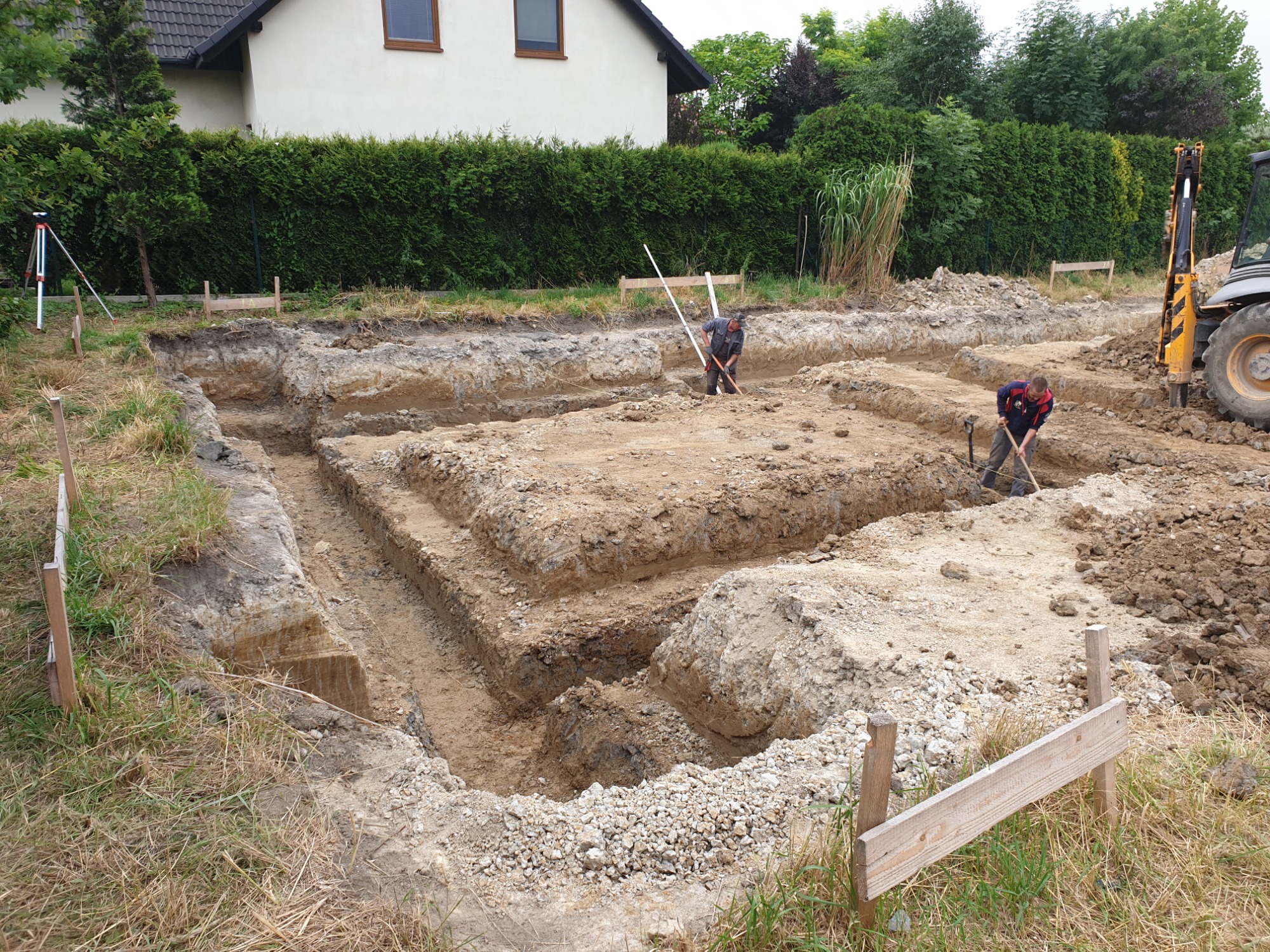 Przygotowanie wykopu pod fundamenty domu murowanego: dwóch robotników kopie łopatami w wyznaczonym obszarze, widoczny żółty traktor i niwelator optyczny.