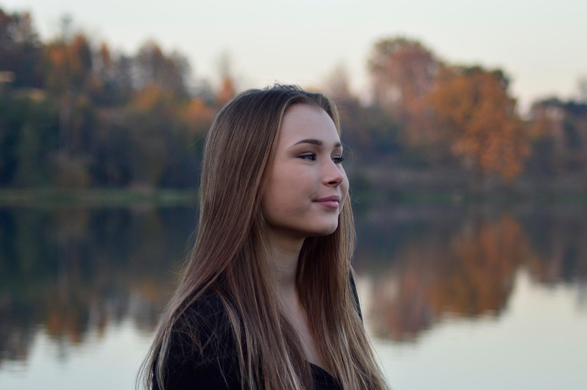 Portret młodej kobiety z długimi, prostymi włosami na tle jesiennego krajobrazu z wodą.