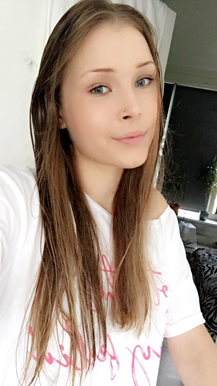 Portret młodej kobiety z kolczykiem w nosie, ubranej w białą koszulkę z różowym napisem, spoglądającej w obiektyw.