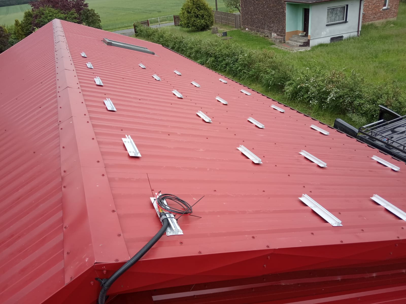 Czerwony blaszany dach z zamontowanymi aluminiowymi profilami montażowymi pod instalację paneli słonecznych, widoczne okablowanie i elementy przyłączeniowe.