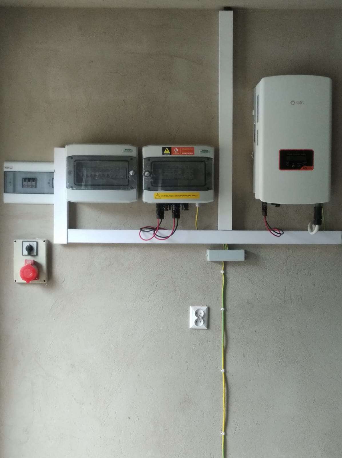 Instalacja elektryczna z inwerterem Solis, zabezpieczeniami w szarych obudowach i wyłącznikiem awaryjnym na ścianie o surowym wykończeniu.