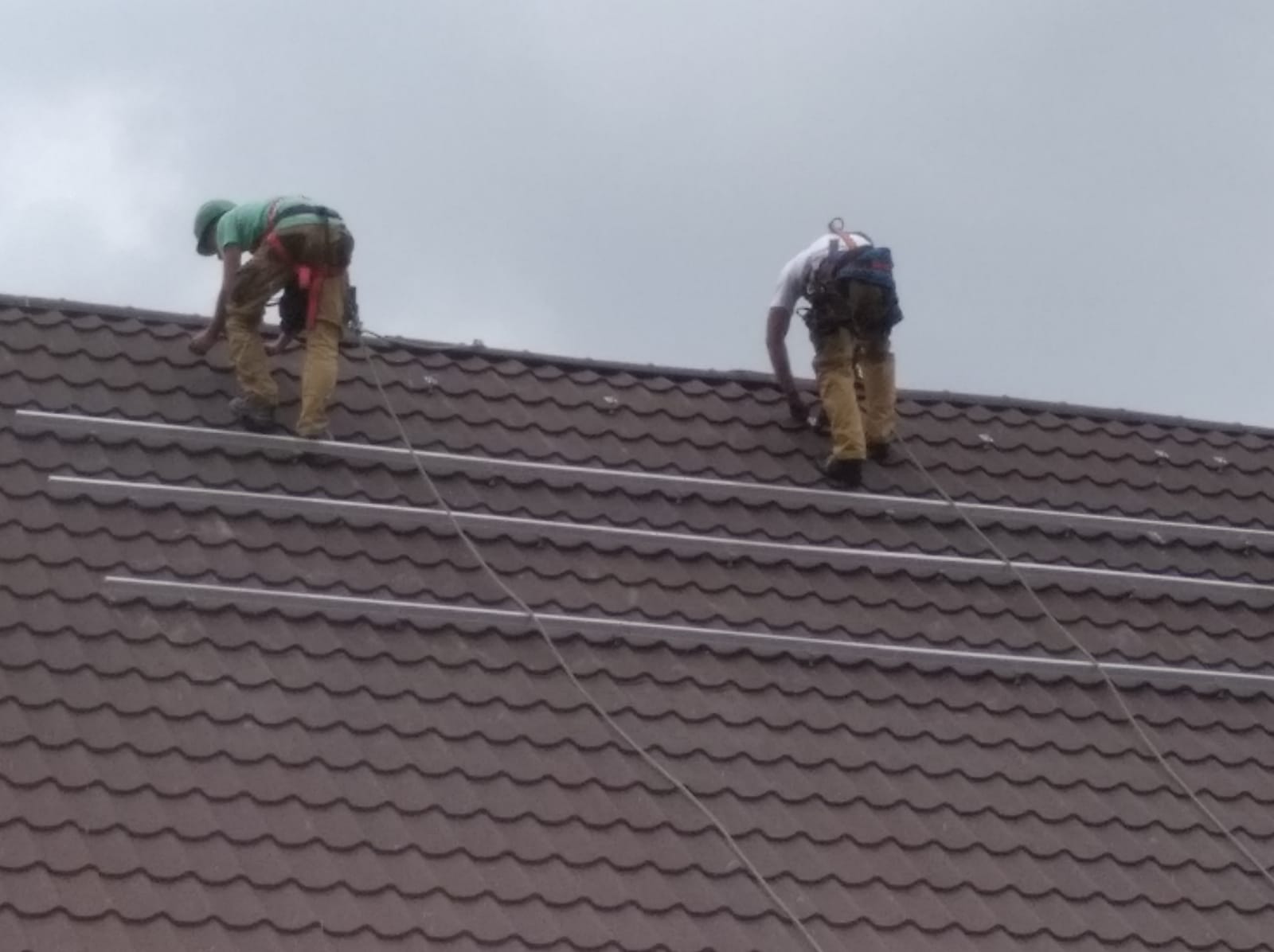 Dwóch monterów w uprzężach zabezpieczających instaluje białe szyny montażowe na brązowym dachu z dachówki ceramicznej pod pochmurnym niebem.