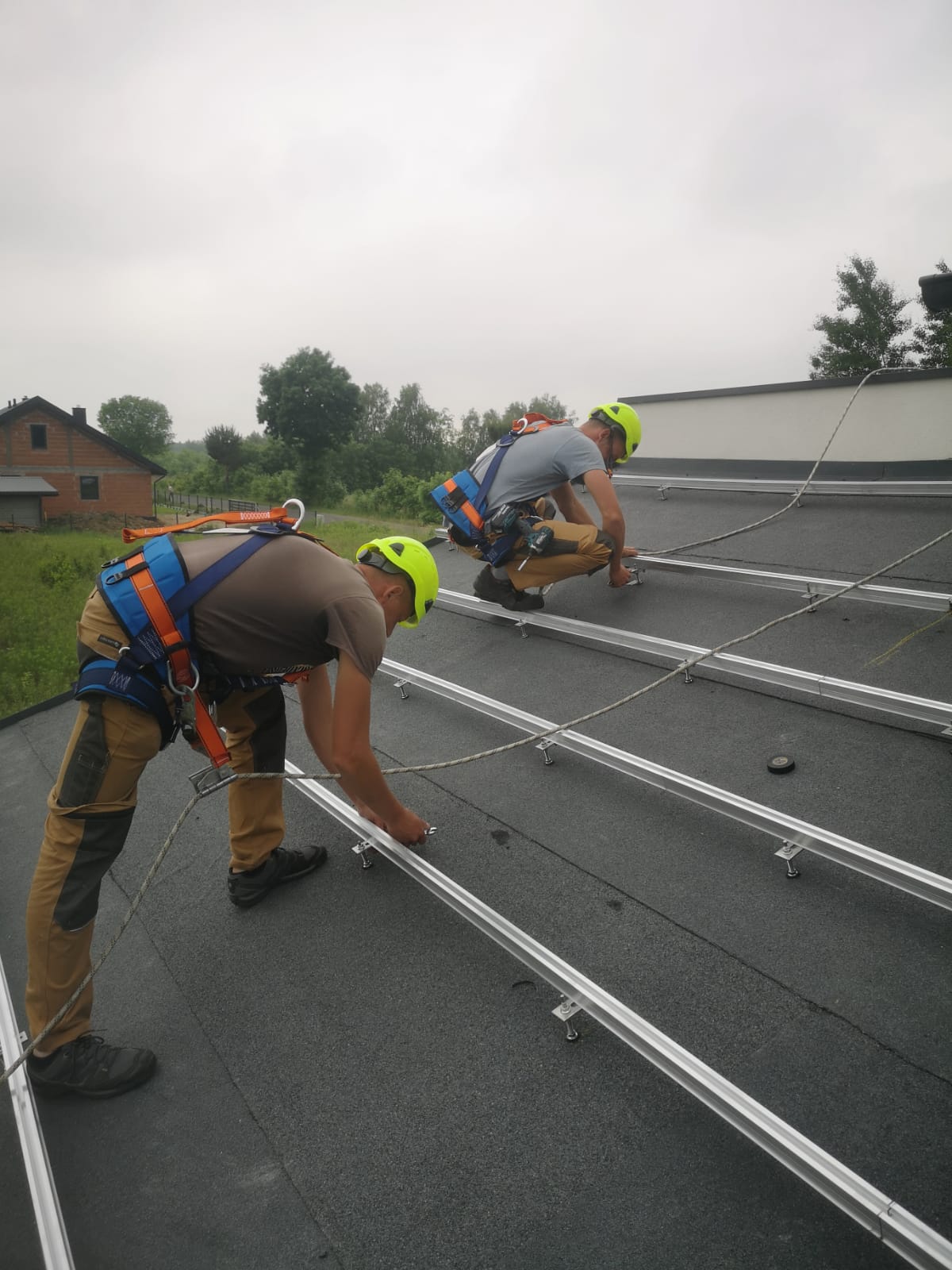Montaż konstrukcji wsporczej pod panele słoneczne na dachu w Popowie. Dwóch monterów w kaskach i uprzężach zabezpieczających przytwierdza aluminiowe profile do pokrycia dachowego.