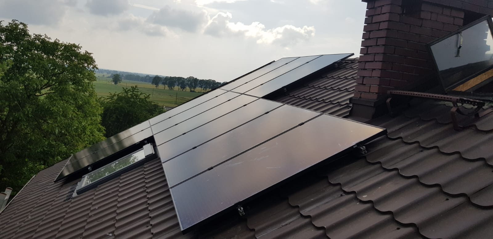 Instalacja fotowoltaiczna na brązowym dachu z dachówki ceramicznej, widok na zielone pola i drzewa w tle, komin z cegły z zamontowanym oknem dachowym.