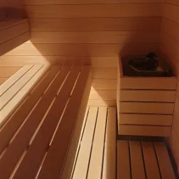 Wnętrze sauny wyłożone jasnym drewnem, z widocznymi ławkami i piecem z kamieniami, oświetlone ciepłym światłem.