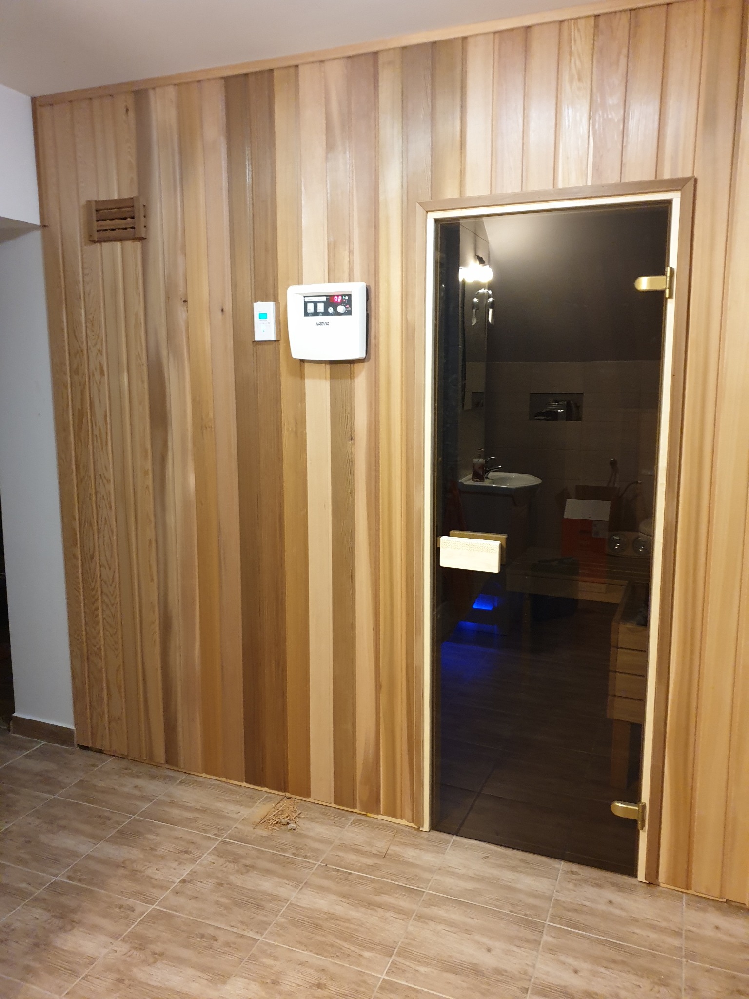 Wnętrze wykończonej sauny z drewnianą boazerią, szklanymi drzwiami, widocznym panelem sterowania i elementami wyposażenia wewnątrz.