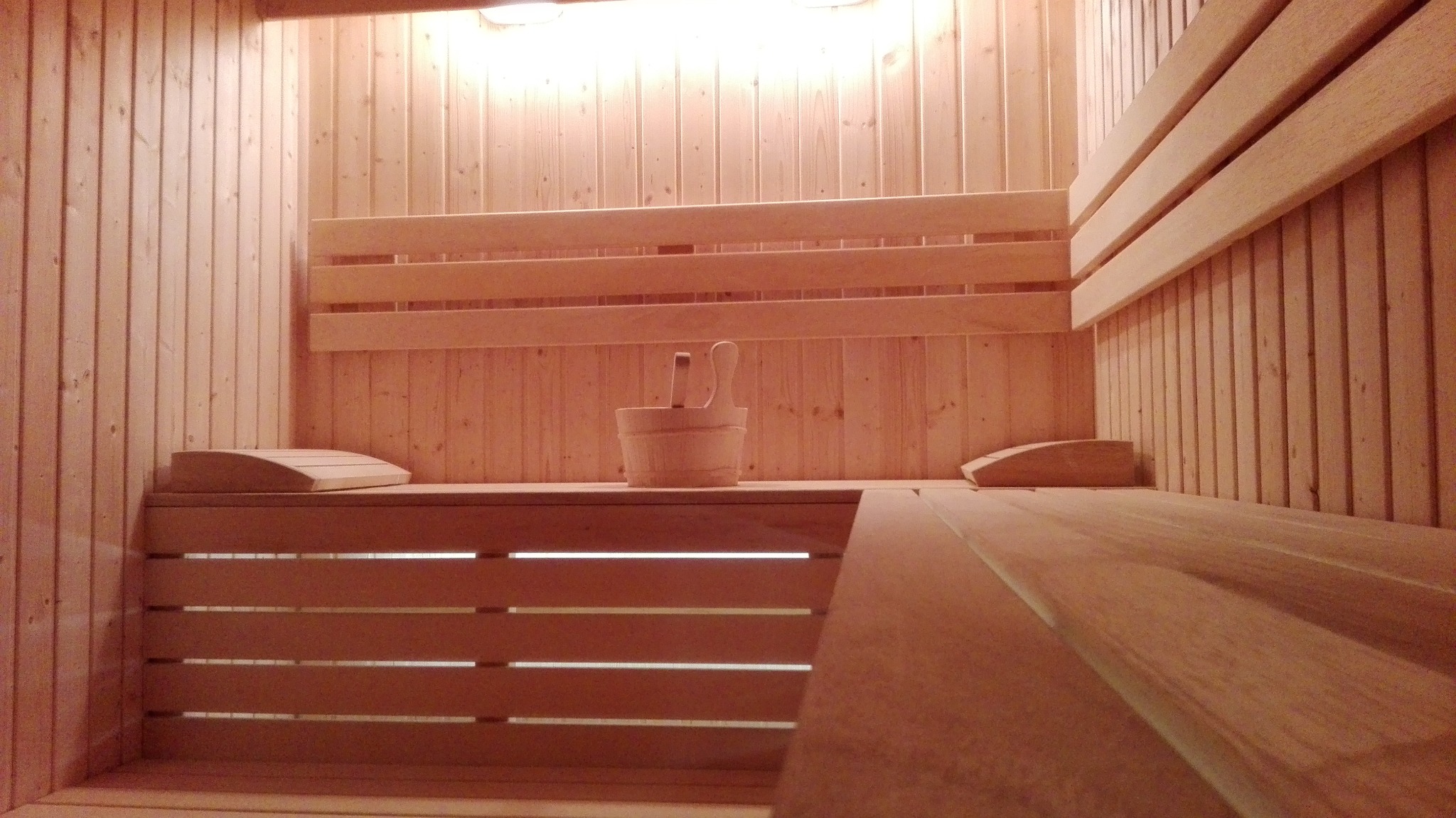 Wnętrze sauny wyłożone jasnym drewnem, z drewnianymi ławkami, oparciami i wiadrem z chochlą, oświetlone górnym światłem.