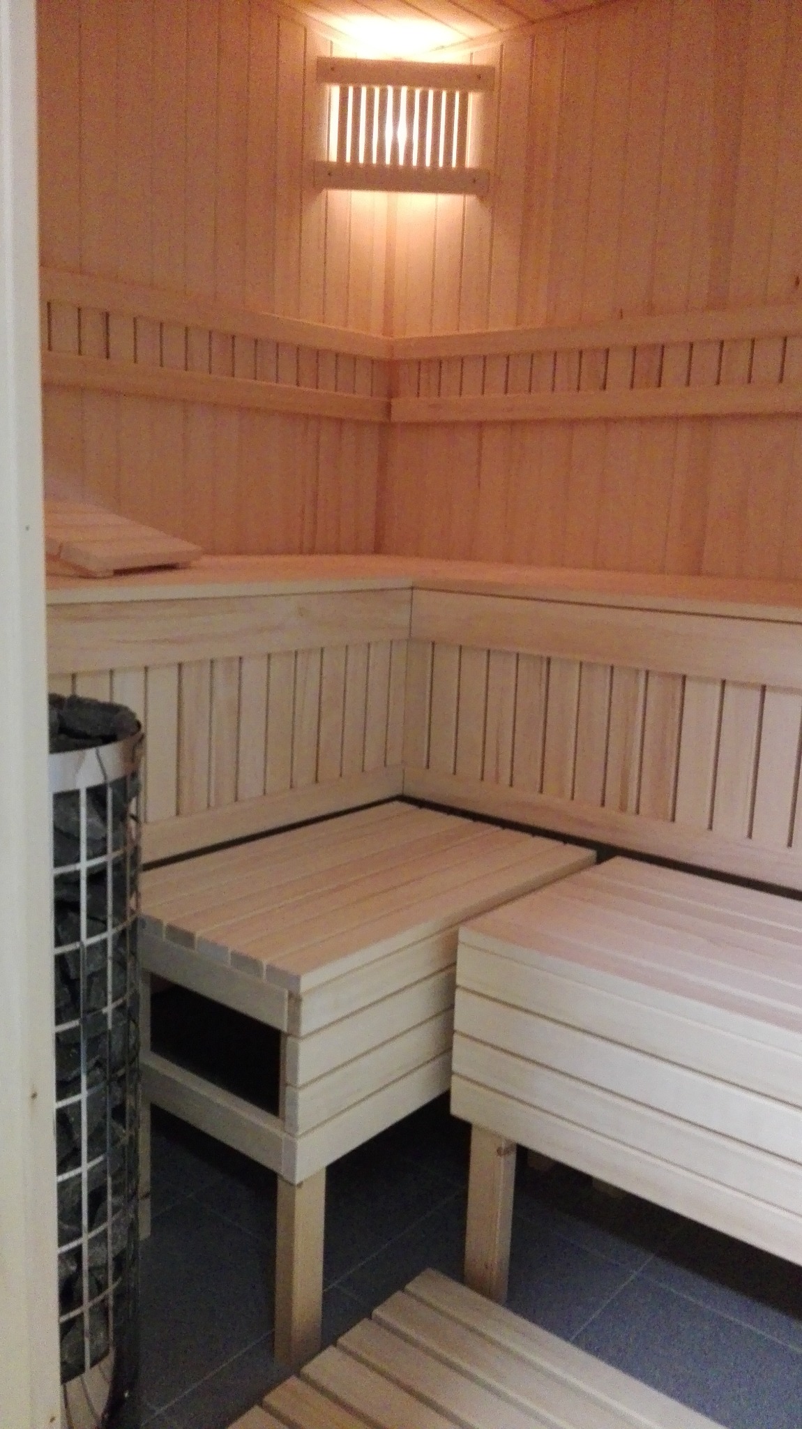 Wnętrze sauny z drewnianymi ławkami, piecem z kamieniami w metalowej siatce i oświetleniem górnym.